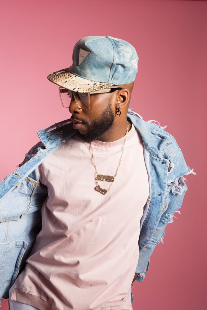 The image depicts a person wearing a light pink t-shirt and a distressed denim jacket. They have a blue baseball cap, and their face is obscured. The background is uniformly pink.